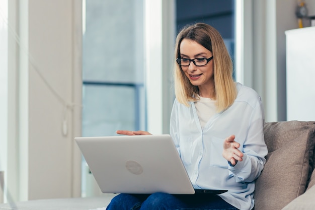 woman in casual clothes sitting on a sofa at home and uses a laptop for video communication 