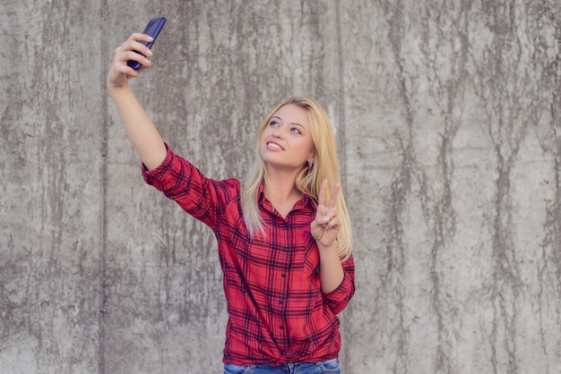 彼女のスマートフォンで自分撮りをタリングし、vサインを見せて笑顔を輝かせるカジュアルな服装の女性