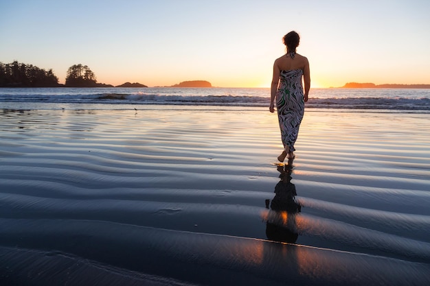 Woman in a casual dress is walking barefoot on a sandy ocean shore towards the water