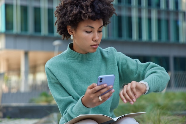 woman in casual jumper checks time on watch waits for someone going to have meeting with friend types text message on cellular poses outside