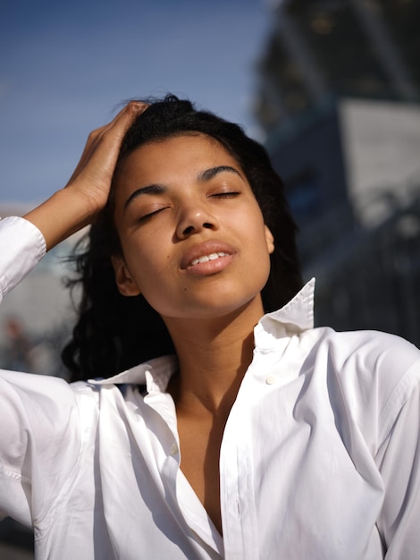 woman in casual white shirt touching her hair while posing with eyes closed outdoors on a warm sunny day. Selective focus