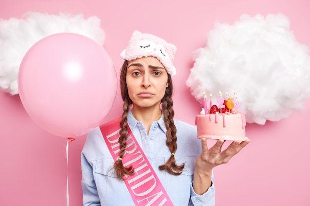 26 歳の誕生日を祝う女性が一人でケーキを持ち、膨らませた風船がピンクに対して悲しそうな表情のポーズをとる
