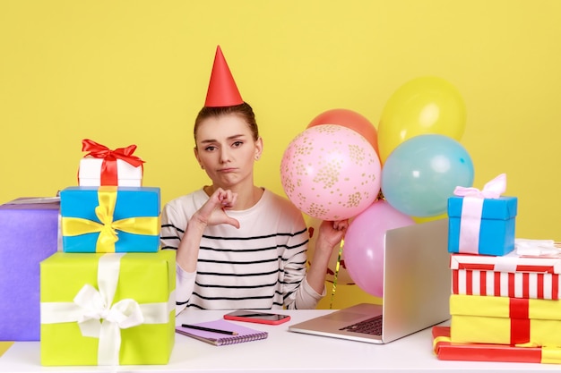 Woman celebrating birthday at workplace among presents and balloons showing thumb down dislike party