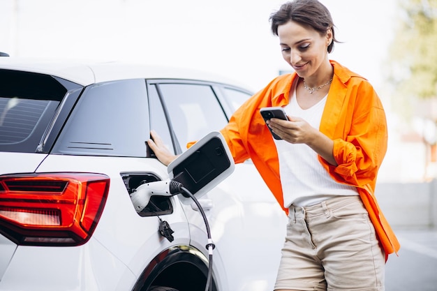 Woman charging electric car at the charging station and using mobile phone