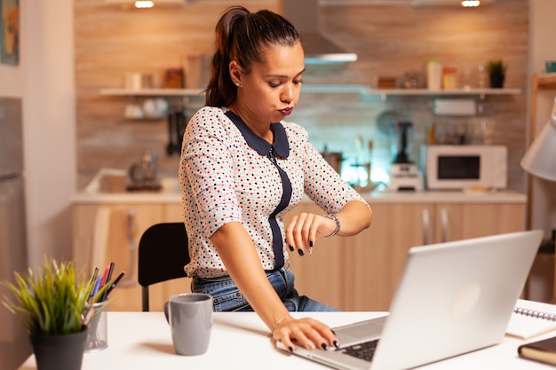 Donna che controlla l'orologio durante il lavoro straordinario nella cucina di casa a mezzanotte. dipendente che utilizza la tecnologia moderna durante le ore notturne facendo gli straordinari per lavoro, affari, impegni, carriera, rete, stile di vita, wireless.