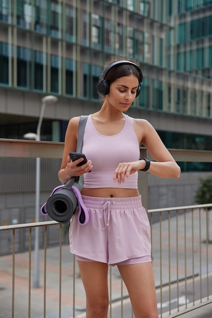  woman checks fitness tracker data or body vitals during training holds modern smartphone with downloaded sport application poses in urban setting prepares for yoga practice