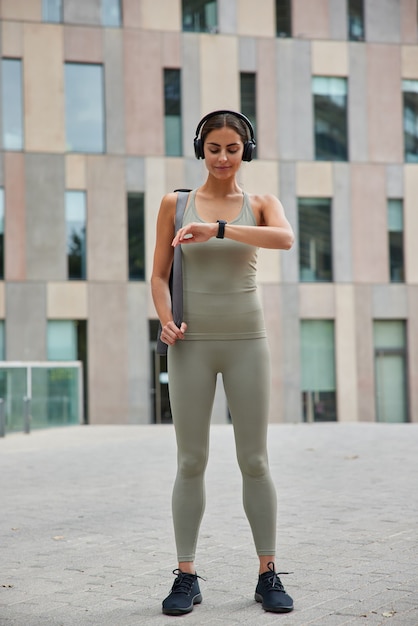 woman checks pulse on smartwatch burned calories after cardio training dressed in activewear carries karemat listens audio track stands outdoor near building