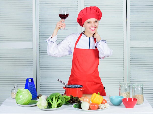 女性シェフがグラスワインを持っています。夕食に添えるワイン。絶妙なディナーのコンセプト。ワインテイスティング。専門家のようにワインと料理を合わせる方法。女の子は帽子とエプロンを着て、飲み物の香りを楽しんでいます。