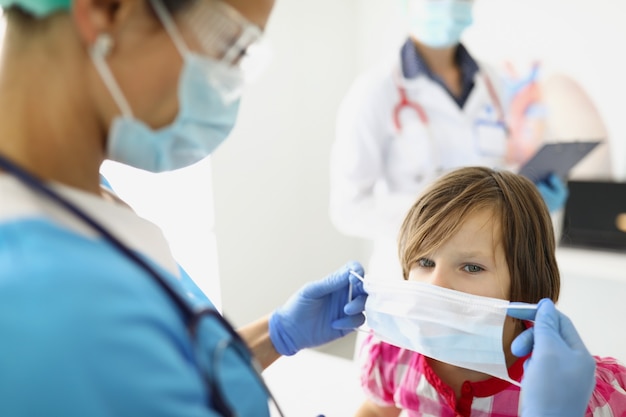 A woman childrens doctor in a mask puts on a protective mask for a girl a child at a doctors