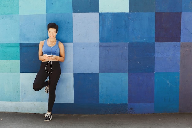 Woman choose music to listen in her mobile phone during workout in city, having rest and leaning at blue painted wall, copy space