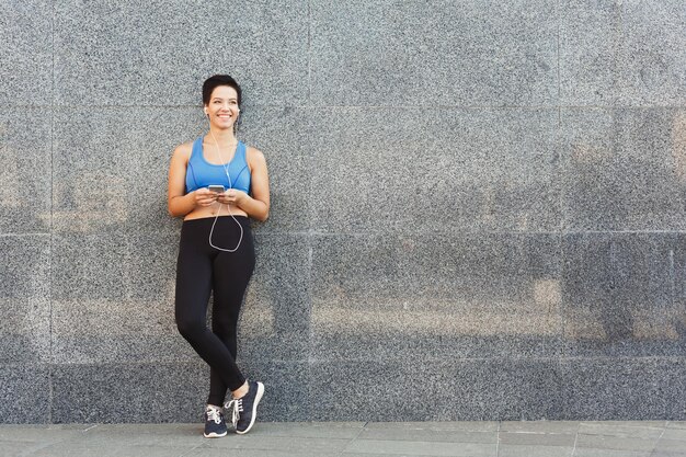 Woman choose music to listen in her mobile phone during workout
in city, having rest and leaning at gray wall, copy space