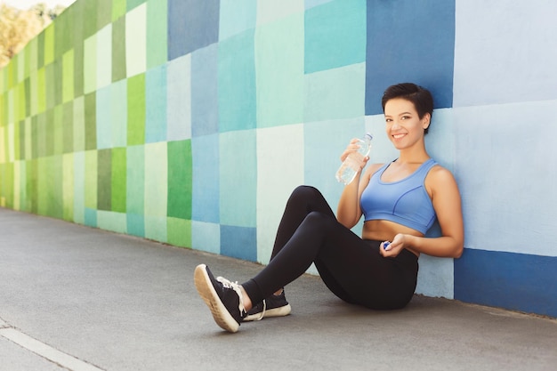 Woman choose music to listen in her mobile phone during workout\
in city, having rest, sitting on floor and leaning at blue painted\
wall, copy space