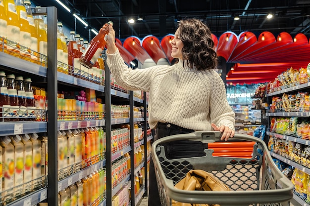 Женщина выбирает сок с полки продуктового магазина, толкая тележку