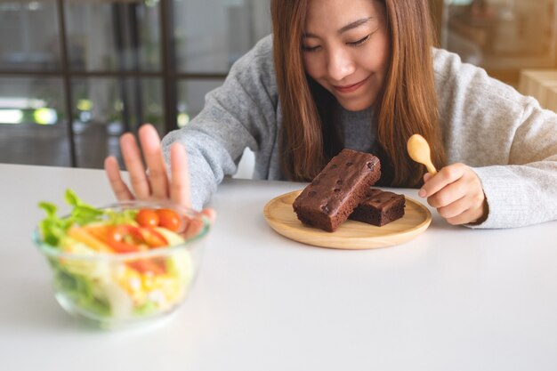 A woman choosing to eat brownie cake and making hand sign to refuse a vegetables salad on the table