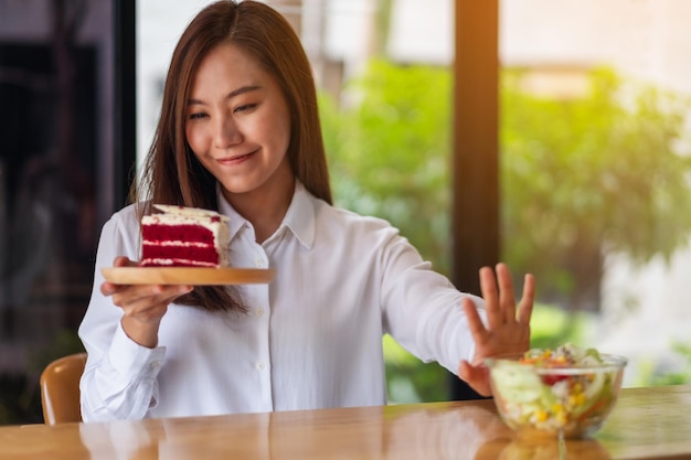 A woman choosing to eat cake and making hand sign to refuse\
vegetables salad on the table