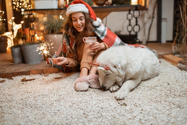 크리스마스 모자를 쓴 여성은 귀여운 강아지와 함께 새해 연휴를 축하하며 집의 아름답게 장식된 테라스에 함께 앉아 있습니다.