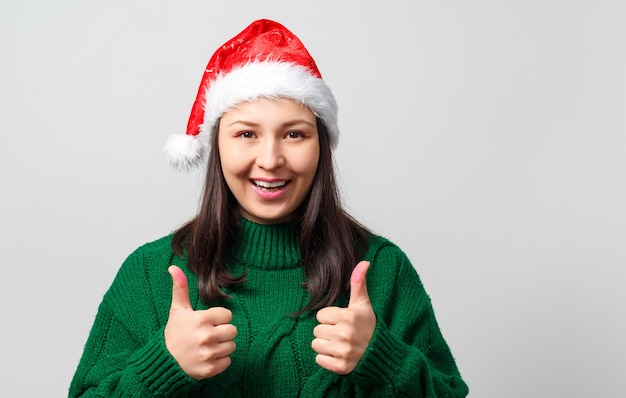제스처를 엄지 손가락을 보여주는 크리스마스 모자에있는 여자. 흰색 배경에.