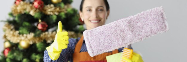 Woman cleaning lady showing thumb up and holding mop near new year tree closeup high quality