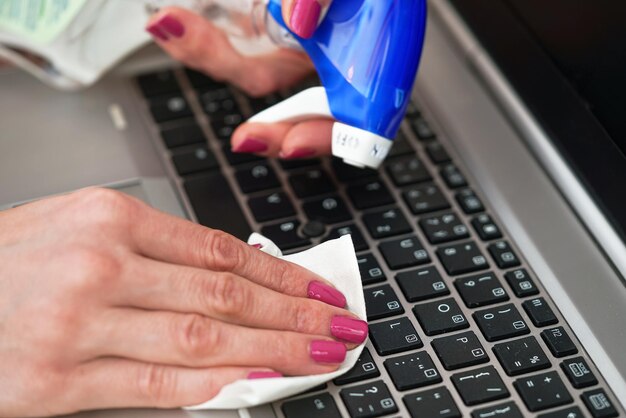 白いティッシュでノートパソコンのキーボードを掃除する女性、ペーパータオルを持つ指のディテール、青いアルコール噴霧器の近く – 消毒のコンセプト。