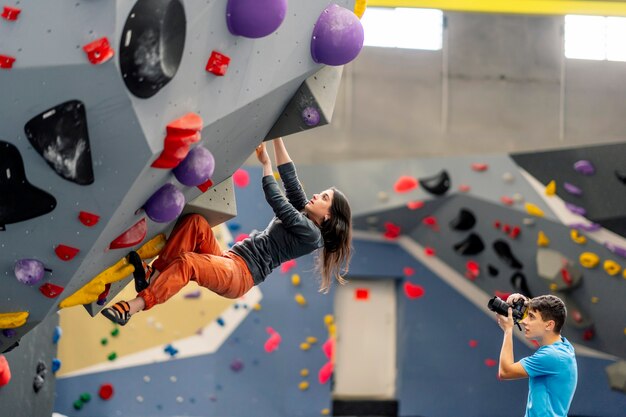 A woman climbing a climbing wall and a man takes a picture of her.