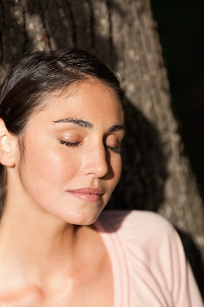 Woman closing her eyes while sitting against a tree as the sun shines on her face