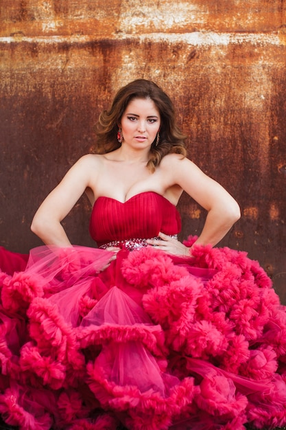 Woman in cloudy red dress is posing over rust wall. Fashion portrait without retouch, only make up