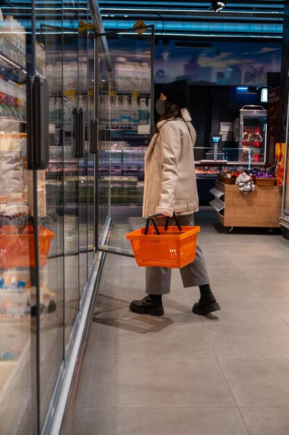 Donna in cappotto e maschera medica al negozio di alimentari che apre la porta del frigorifero