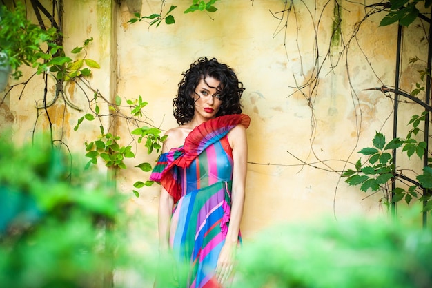 A woman in a colorful dress stands in front of a wall with vines growing on it.