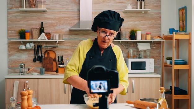 Woman communicating with subscribers through video camera while kneading dough. Retired blogger chef influencer using internet technology shooting blogging on social media with digital equipment