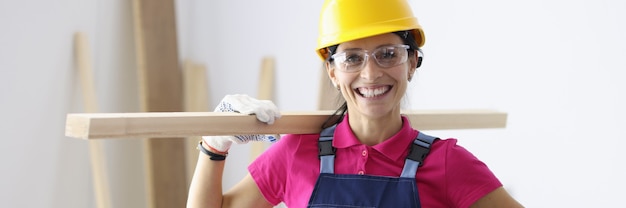 Woman in construction helmet and overalls holding wooden plank on shoulder in workshop. Carpentry work concept