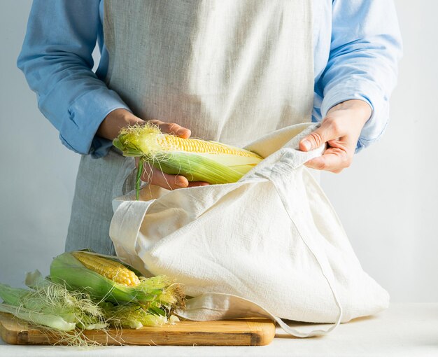 Woman cook in a cotton apron holds a string bag with ears of fresh ripe corn organic food concept kinfolk concept