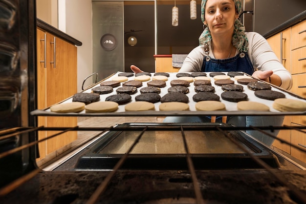 Foto cuoco che introduce una teglia con coperchi di pasta di cacao all'interno del forno per la preparazione della ricetta casalinga degli alfajores argentini. concetto di cibo regionale e tradizionale.