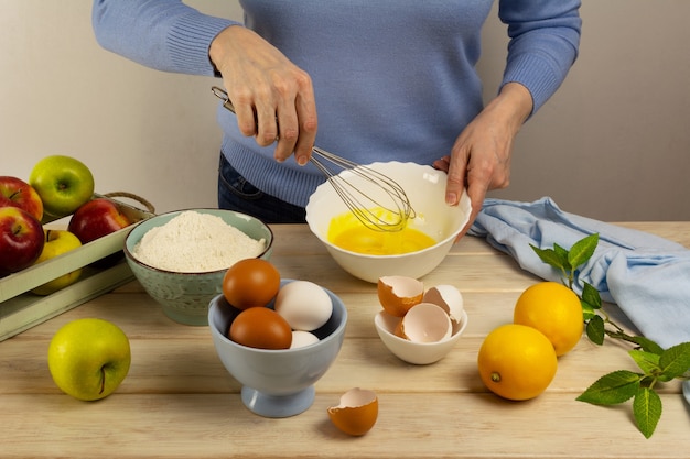 Woman cooking apple pie dough in the white bowl and fruits on the wooden table