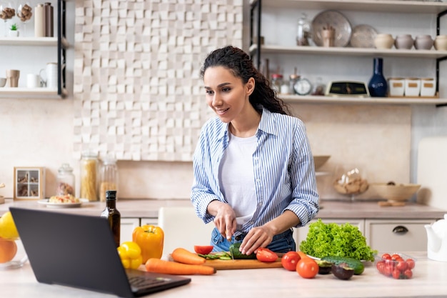 Woman cooking fresh organic salad cutting cucumber and looking at laptop on countertop preparing