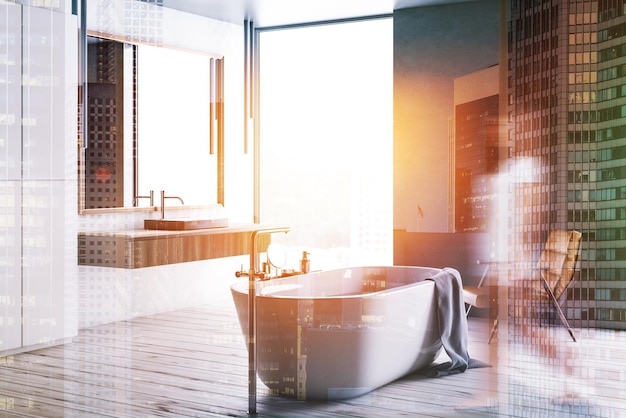 Женщина в углу современной ванной комнаты с серыми и белыми деревянными стенами, окном на чердаке, белой ванной и серой раковиной на деревянном прилавке с большим зеркалом над ним.