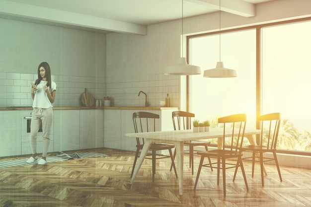 Photo woman in corner of modern kitchen with white walls, wooden floor, large window, white countertops and wooden table with black chairs. toned image double exposure