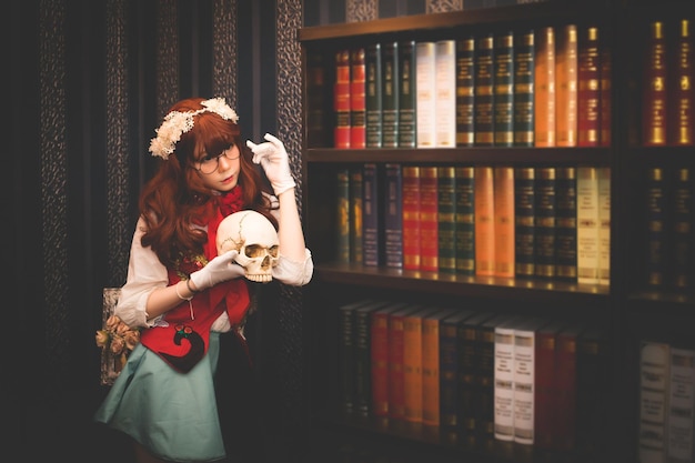 Photo woman in costume holding skull while standing by books in shelves
