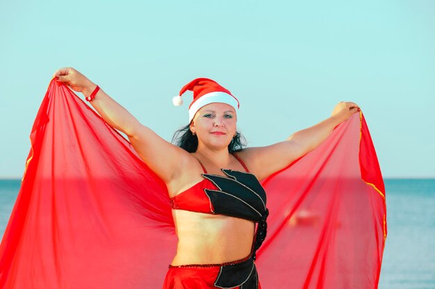 A woman in a costume for oriental dances in a Santa hat against the background of the sea is dancing with a scarf.