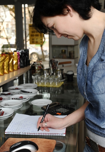 Woman behind the counter of a cafe is taking notes in a notebook.