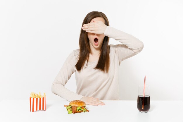 Фото Женщина закрыла глаза рукой за столом с гамбургером, картофелем-фри, колой в стеклянной бутылке, изолированной на белом фоне. правильное питание или классический американский фастфуд. рекламная площадка с копией пространства.