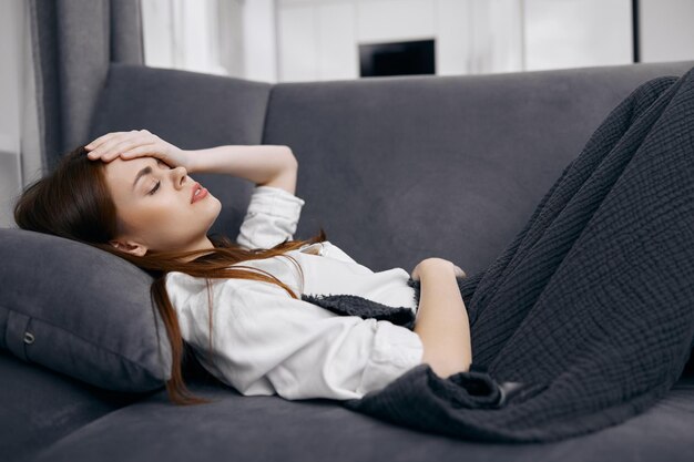 Фото Женщина, покрытая одеялом, лежит на диване, положив руку на голову, плохо себя чувствует
