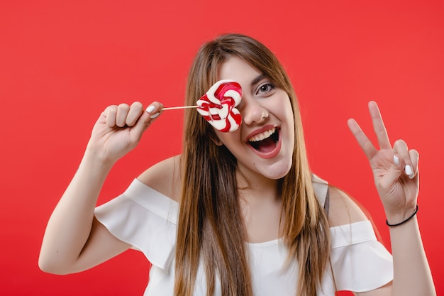 Woman covering eye with heart shaped lollypop candy wearing white blouse isolated on red wall