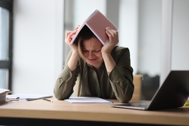 Woman covers head with book feeling tired and powerless female employee sits at table near
