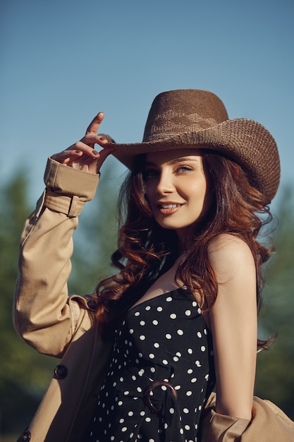 woman in a cowboy hat resting in the village near the stables, portrait of a woman in the sun, rustic style. Portrait of a sexy brunette in nature. Perfect makeup, natural cosmetics