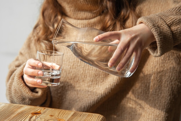 A woman in a cozy sweater pours water into a glass from a glass decanter, the concept of maintaining water balance.