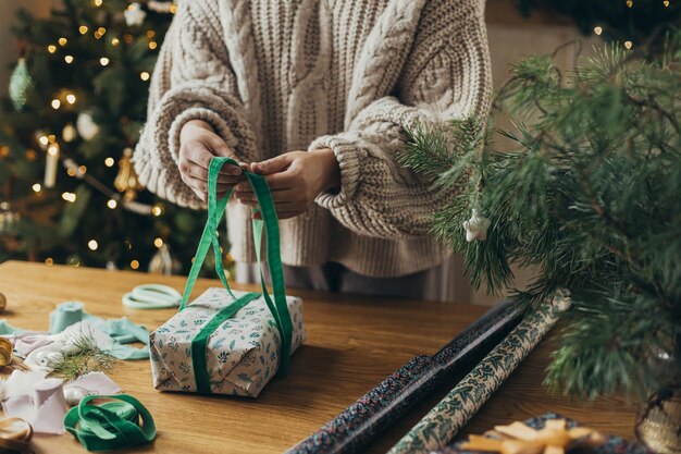 Woman in cozy sweater wrapping stylish christmas gift in festive wrapping paper with ribbons vintage ornaments bows on wooden table atmospheric winter holidays wrapping christmas present