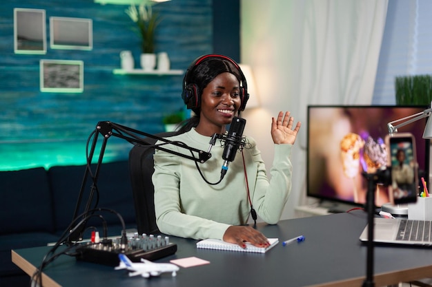 Woman creator sitting at table in streaming studio greeting subscribers while recording video content for youtube channel. Social media influencer filming vlog talking into production micophone