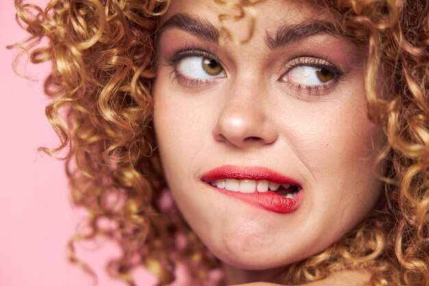 Woman Curly hair grimace bites her lip with a look towards the pink background bright makeup closeup