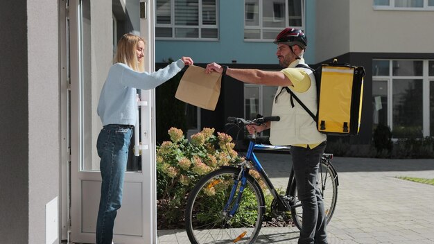 Woman customer open door to a food delivery man wearing yellow thermal backpack on a bike and recieves order or package with food Food delivery concept