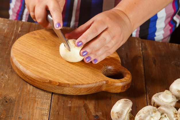 A woman cuts champignon mushrooms with a knife on a round wooden board